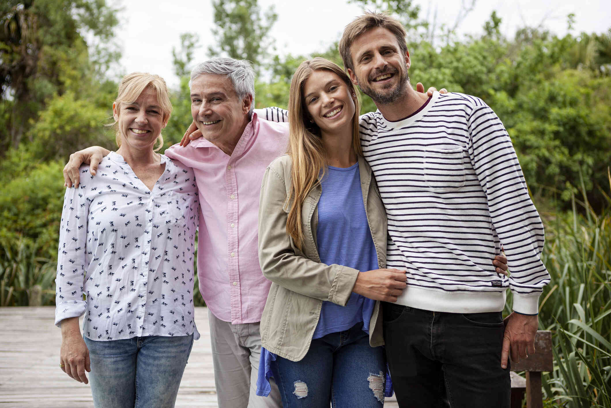 A family of four adults embrace each other happily while standing outside on a sunny  day and smiling at the camera.